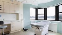 A medical exam room in the new CUIMC Student Health Center is shown. It features a stool, an exam table, with pale wood cabinets, light blue walls and a large bay window with a view of the Hudson River.